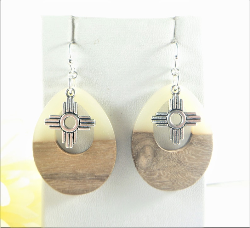 Puede incluir: Un par de pendientes de madera con un dije de plata en forma del s&iacute;mbolo Zia. Los pendientes son de forma ovalada y tienen un patr&oacute;n de veta de madera marr&oacute;n claro y blanco.
