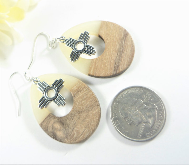 Puede incluir: Un par de pendientes de madera con un dije de plata con el s&iacute;mbolo Zia. Los pendientes son de forma ovalada y tienen madera marr&oacute;n claro en un lado y resina blanca en el otro. Los pendientes son aproximadamente del tama&ntilde;o de una moneda de 25 centavos.