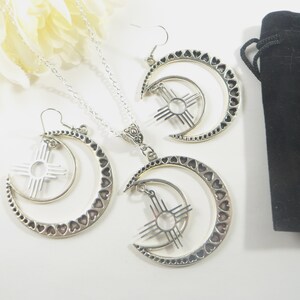 May include: A silver-toned jewelry set featuring a necklace and earrings. The necklace has a pendant with a crescent moon design, adorned with heart cutouts, and a sun symbol. The earrings mirror the pendant design. A black velvet pouch is also included.