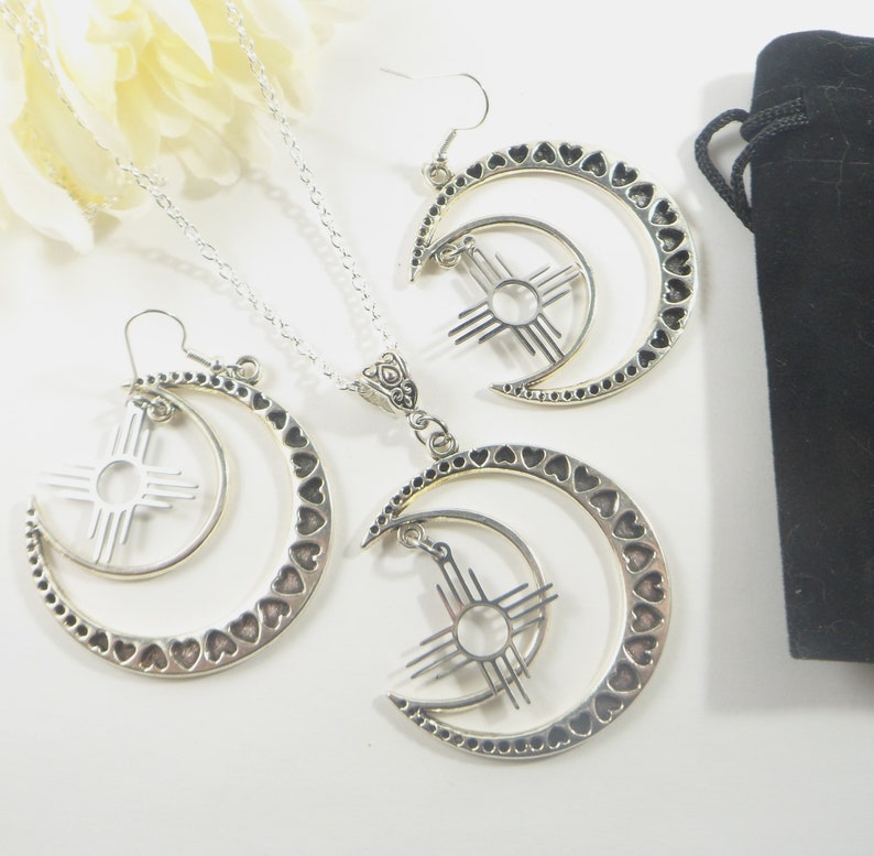 May include: A silver-toned jewelry set featuring a necklace and earrings. The necklace has a pendant with a crescent moon design, adorned with heart cutouts, and a sun symbol. The earrings mirror the pendant design. A black velvet pouch is also included.