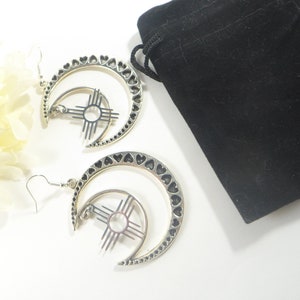 May include: A pair of silver-toned earrings featuring a crescent moon design with heart-shaped cutouts along the edge. Each earring has a sun symbol in the center. A black velvet pouch is in the background.