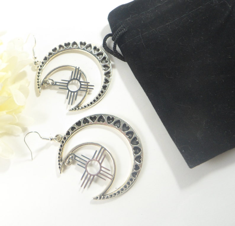 May include: A pair of silver-toned earrings featuring a crescent moon design with heart-shaped cutouts along the edge. Each earring has a sun symbol in the center. A black velvet pouch is in the background.