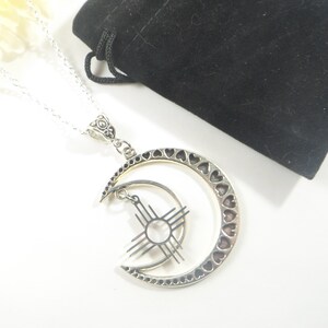 May include: Silver-toned necklace featuring a crescent moon pendant with heart cutouts and a smaller inner moon with a sunburst design. The necklace is displayed on a white surface with a black velvet pouch in the background.
