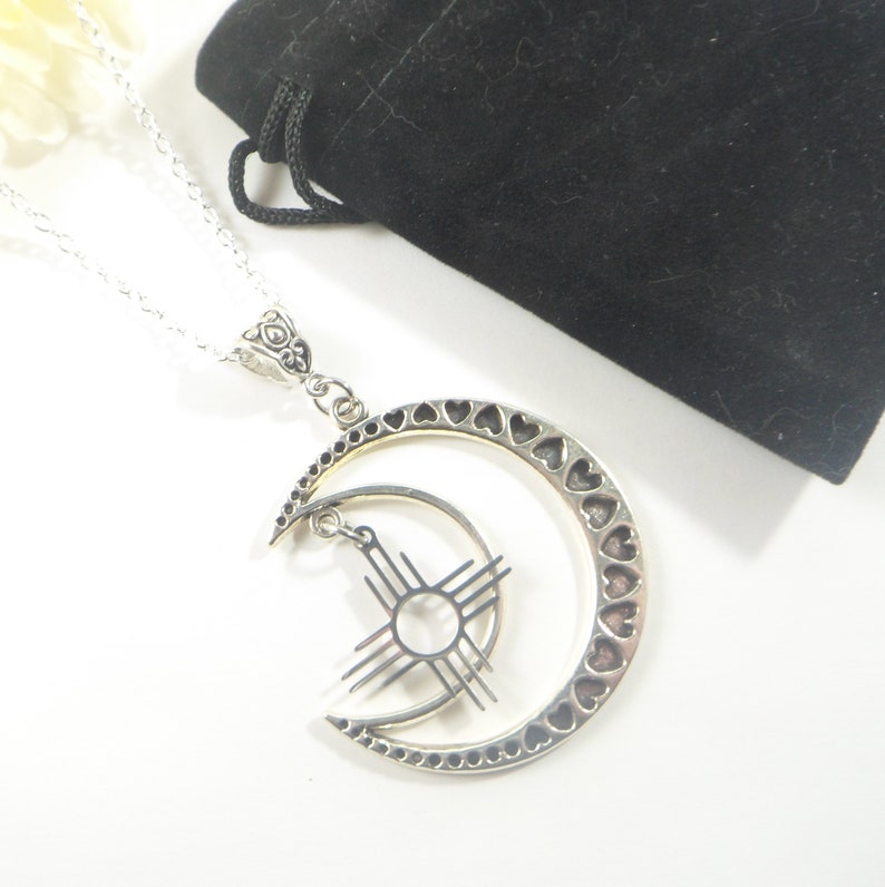May include: Silver-toned necklace featuring a crescent moon pendant with heart cutouts and a smaller inner moon with a sunburst design. The necklace is displayed on a white surface with a black velvet pouch in the background.