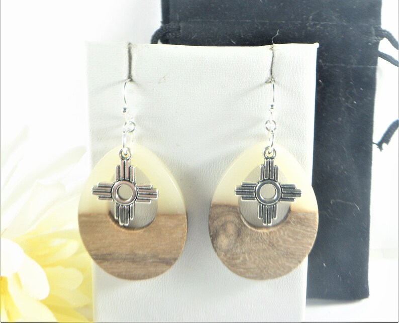 Puede incluir: Un par de pendientes de madera con un dije de plata con el s&iacute;mbolo Zia. Los pendientes son de forma ovalada y tienen un patr&oacute;n de veta de madera blanca y marr&oacute;n.