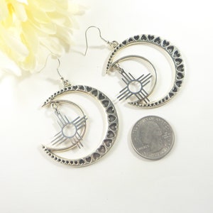May include: Silver-toned earrings featuring a crescent moon design with heart cutouts and a sun symbol. The earrings have a hook closure and are displayed on a white surface with a quarter for size reference. A yellow flower is in the background.