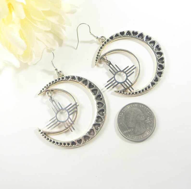 May include: Silver-toned earrings featuring a crescent moon design with heart cutouts and a sun symbol. The earrings have a hook closure and are displayed on a white surface with a quarter for size reference. A yellow flower is in the background.