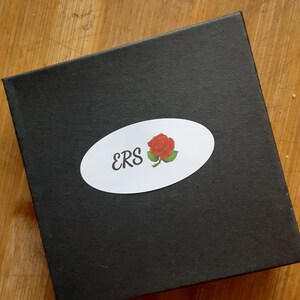 Puede incluir: Caja negra con una pegatina ovalada blanca con el texto "ERS" y una ilustraci&oacute;n de rosa roja.