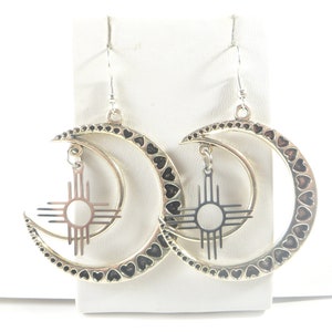 May include: Silver-toned earrings featuring a crescent moon design with heart-shaped cutouts along the edges. Each earring has a dangling sun symbol pendant. The earrings are displayed on a white surface.