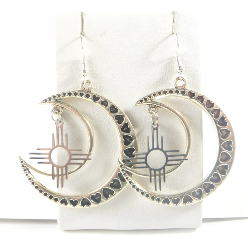 May include: Silver-toned earrings featuring a crescent moon design with heart-shaped cutouts along the edges. Each earring has a dangling sun symbol pendant. The earrings are displayed on a white surface.