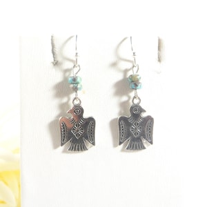May include: A pair of silver-toned earrings featuring a stylized bird design. Each earring has a turquoise bead accent and a decorative bird pendant with etched details. The earrings hang from silver-colored ear wires.