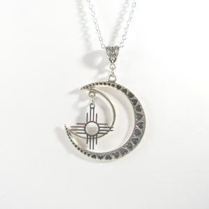 May include: Silver-toned necklace featuring a crescent moon pendant with heart-shaped cutouts. A smaller pendant with a sun symbol hangs within the moon. The necklace has a delicate chain and ornate bail.