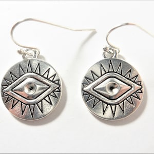 May include: A pair of silver earrings with a circular design featuring a stylized eye with a sunburst pattern around it.