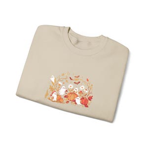 Può includere: Una felpa girocollo color beige con una stampa grafica centrale raffigurante fantasmi bianchi, zucche arancioni, funghi rossi, margherite bianche e piccoli pipistrelli marroni circondati da foglie e viticci autunnali nei toni dell'arancio.
