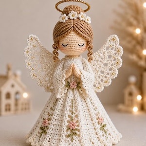 May include: A crocheted angel figurine with light brown hair, a gold halo, and white wings accented with small gold beads. The angel wears a white dress featuring embroidered pink roses with green stems and leaves, with hands clasped in a prayer position.