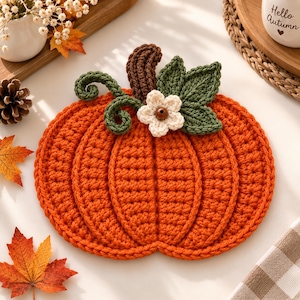 Puede incluir: Un posavasos de ganchillo naranja con forma de calabaza redonda, que presenta un tallo marrón, dos zarcillos verdes, dos hojas verdes y una pequeña flor blanca con el centro marrón. La calabaza tiene una textura acanalada.
