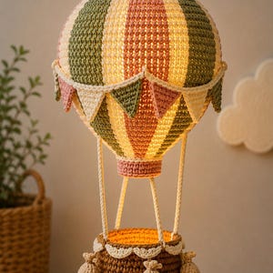 May include: A crocheted hot air balloon lamp featuring vertical stripes in sage green, cream, and dusty pink. The balloon is adorned with a small triangular bunting, while the brown basket below is decorated with three small crocheted pouches.