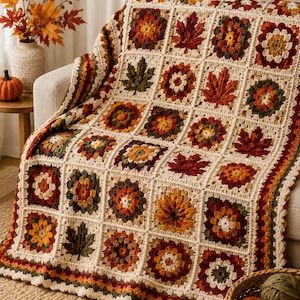 May include: A crochet blanket featuring a grid of square motifs in autumn colors including cream, rust orange, deep red, and forest green. The squares alternate between floral-style patterns and detailed maple leaf designs with a scalloped border.