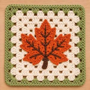 May include: A square crochet granny square featuring a central orange maple leaf with dark brown veins and a stem. The leaf is centered on a white background, framed by a border of sage green stitches, creating a textured, handcrafted textile piece.
