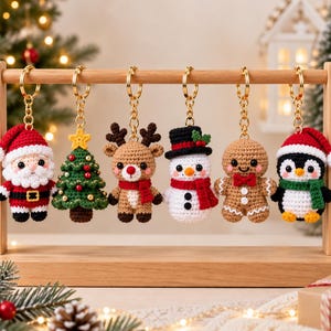 Op de afbeelding: Een set van zes gehaakte kerst-sleutelhangers hangend aan een houten standaard. De figuren omvatten de Kerstman, een versierde groene kerstboom, een bruin rendier, een sneeuwpop met een zwarte hoed, een peperkoekmannetje en een pinguïn met een rode muts.