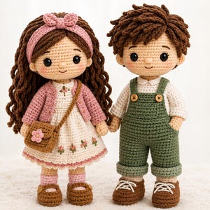 May include: Two handmade crochet dolls standing side by side. The left doll has long brown hair, a pink headband, a pink cardigan, and a cream dress with floral embroidery. The right doll has short brown hair, a white shirt, and green overalls.