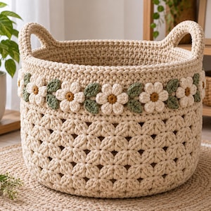 Peut inclure: Un panier rond en crochet de couleur crème doté de deux anses intégrées. La partie supérieure est ornée d'une bande décorative de fleurs blanches à cœur jaune et de feuilles vertes, tandis que le bas présente un motif en forme d'étoile.