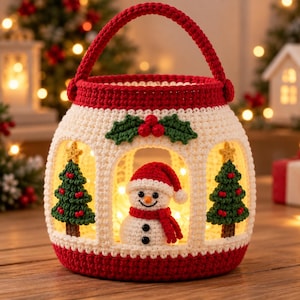 May include: A crocheted lantern basket featuring a red handle and base with a cream-colored body. The design includes two green Christmas trees, a central snowman wearing a red hat and scarf, and a holly berry accent at the top.