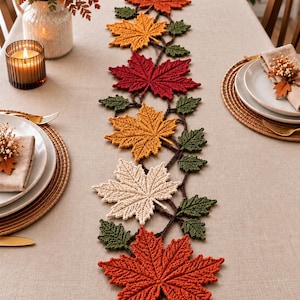 May include: A crochet table runner featuring a series of interconnected maple leaves in shades of orange, yellow, red, and cream, accented with smaller dark green leaves. The runner is arranged in a vertical line down the center of a table.
