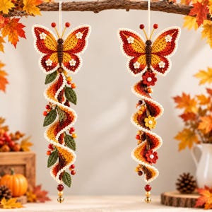 Può includere: Due decorazioni a farfalla all'uncinetto da appendere con ali nei toni del rosso, arancione e giallo con dettagli bianchi. Ogni farfalla è fissata a una coda a spirale multicolore decorata con piccole foglie verdi, bacche rosse e fiori.
