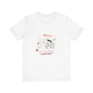 Puede incluir: Una camiseta blanca de manga corta con un gráfico central de un marco de fotos rectangular que contiene un dibujo lineal en blanco y negro de dos personas. El diseño incluye pequeños corazones rosas, un acento de cinta rosa y el texto Love You Forever.