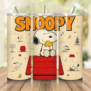 May include: A cylindrical stainless steel tumbler with a cream-coloured background featuring the word SNOOPY in large orange letters. The design includes illustrations of Snoopy and Woodstock, a red doghouse, musical notes, and a typewriter.