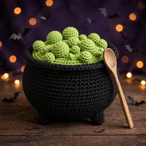 May include: A black crocheted cauldron filled with various sizes of light green crocheted spheres, accompanied by a single wooden spoon resting against the side of the pot. The item features a textured, handmade yarn construction.