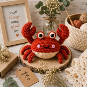 May include: A crocheted crab toy with a bright red upper body, a cream-coloured underbelly, two raised claws, and two large black eyes. The crab features small pink stitched cheeks and a simple black smile, resting on a round wooden slice.
