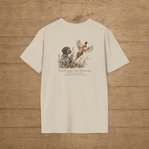 May include: A beige short-sleeve t-shirt featuring a graphic of a brown dog sitting in tall grass with a pheasant flying above. The text reads: Good Dogs. Bad Shooting. FIELD & FLEDGE. Midwest Outdoor Goods. The design is centered on the back.