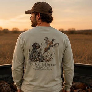 May include: A beige long-sleeve shirt featuring a graphic on the back of a hunting dog sitting in tall grass with a pheasant in flight. The text reads: Good Dogs. Bad Shooting. FIELD & FLEDGE. Midwest Outdoor Goods. The shirt is worn with a brown cap.