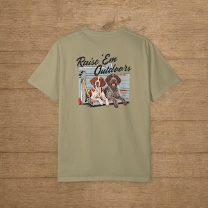 May include: A sage green short-sleeve t-shirt featuring a graphic on the back. The illustration depicts two dogs sitting in the back of a light blue truck bed with the text Raise 'Em Outdoors printed in black script above the canine subjects.