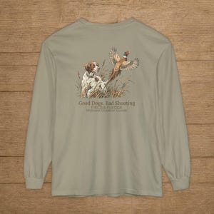 May include: A sage green long-sleeve t-shirt featuring a graphic of a brown and white dog in tall grass watching a pheasant take flight. The text reads: Good Dogs. Bad Shooting. FIELD & FLEDGE. Midwest Outdoor Goods.