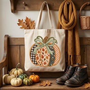 May include: A cream-colored canvas tote bag featuring a large central graphic of a pumpkin decorated with various patterns including black and white buffalo check, autumn leaves, and stripes, tied with a light green ribbon bow at the top.