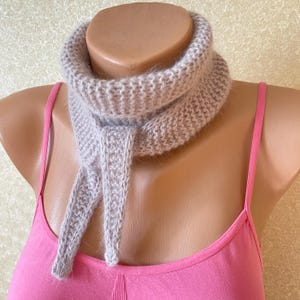 May include: A light beige knitted neck warmer with a fuzzy texture and a ribbed pattern. The accessory features a high collar design that wraps around the neck, with two pointed, dangling ends hanging down the front of the garment.