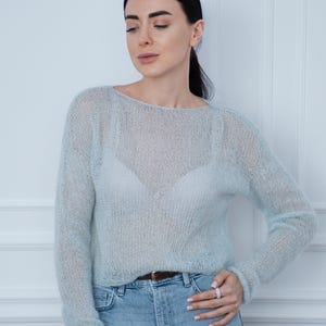 May include: A light blue, long-sleeved, sheer knit sweater with a wide boat neckline. The sweater is crafted from a fuzzy, lightweight yarn with a loose, open-stitch construction, worn over a matching light blue camisole and light blue denim jeans.
