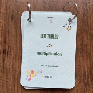 Könnte beinhalten: Ein Set hellblauer Lernkarten, die mit zwei silbernen Metallringen zusammengehalten werden. Die Vorderseite zeigt den Text "LES TABLES De multiplication" in Dunkelgrün, "Aide à la mémorisation" und "De 1 à 10" sowie bunte Zahlen und Sternsymbole.