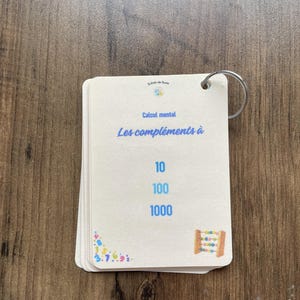 May include: A stack of off-white educational flashcards held together by a silver metal ring. The top card features the blue text "Calcul mental" and "Les compléments à" above the numbers 10, 100, and 1000, with a small abacus illustration.