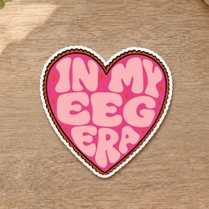 May include: A heart-shaped sticker with a bright pink background and a dark brown scalloped border. The text IN MY EEG ERA is written in large, light pink, wavy bubble letters. Small orange diamond shapes are scattered between the words.