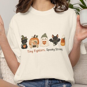 Puede incluir: Una camiseta color hueso con un gráfico de un gato negro, una calabaza, un fantasma, un murciélago y un dulce, cada uno con un icono de corazón. Debajo del gráfico, el texto dice Tiny Fighters, Spooky Strong. Tiene mangas cortas.