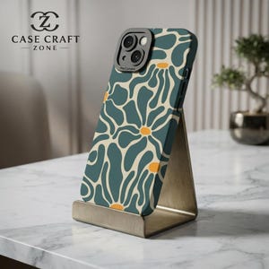 Puede incluir: Una funda para smartphone con un patrón floral abstracto en azul verdoso oscuro y crema con pequeños acentos circulares amarillos. La funda tiene un recorte negro para el módulo de cámara y se muestra en un soporte metálico. El texto CASE CRAFT ZONE es visible.