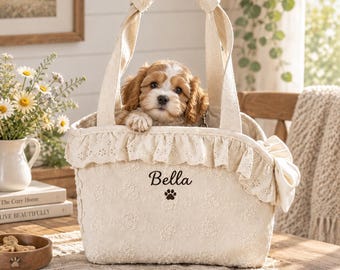 May include: A cream-coloured fabric pet carrier bag featuring a ruffled eyelet trim, two sturdy handles, and a side bow. The front is embroidered with the name Bella above a small paw print icon. A small brown and white puppy sits inside.