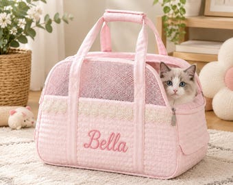 May include: A light pink quilted pet carrier featuring a mesh top panel, a decorative cream-coloured lace trim, and the name Bella embroidered in pink script on the front. A fluffy white and grey kitten is visible inside the carrier.