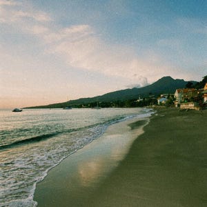 Peut inclure: Un paysage côtier avec une plage de sable sombre rencontrant les vagues calmes de l'océan au coucher du soleil. Au loin, une chaîne de montagnes s'élève derrière une rangée de bâtiments le long du rivage, avec quelques petits bateaux sur l'eau.