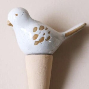 May include: A bottle stopper featuring a white ceramic bird with brown speckled wing details and a tan beak. The bird is mounted on a tapered, light-coloured wooden base. The ceramic surface has a glossy finish with visible dark speckles throughout.