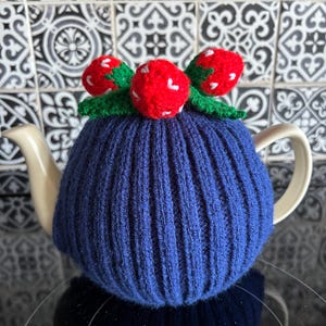 May include: A blue knitted tea cozy with a ribbed design, topped with three red strawberries with white accents and green leaves. The cozy is designed to fit over a white teapot, adding a decorative touch to tea time.